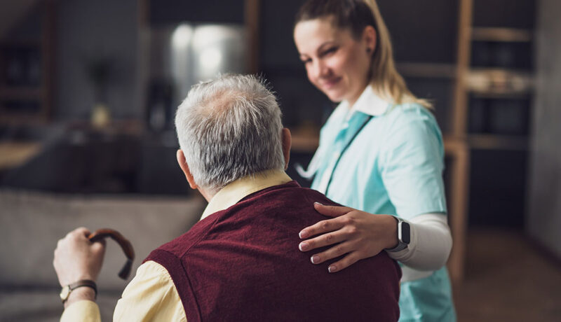 A Caregiver Smiling With Her Hand on a Senior Man’s Back Sitting Down Family Owned Assisted Living